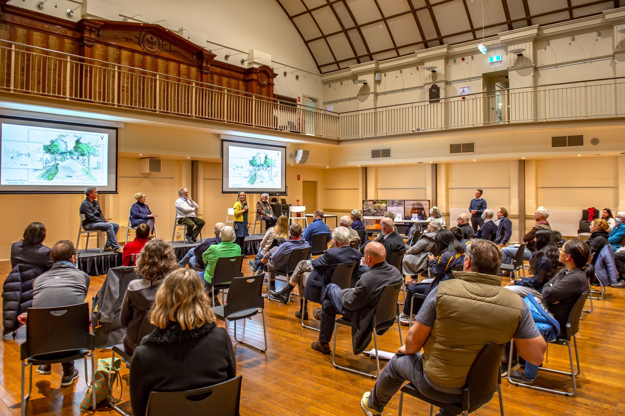 Community meeting in a hall with people seated facing presenters and large display screens.